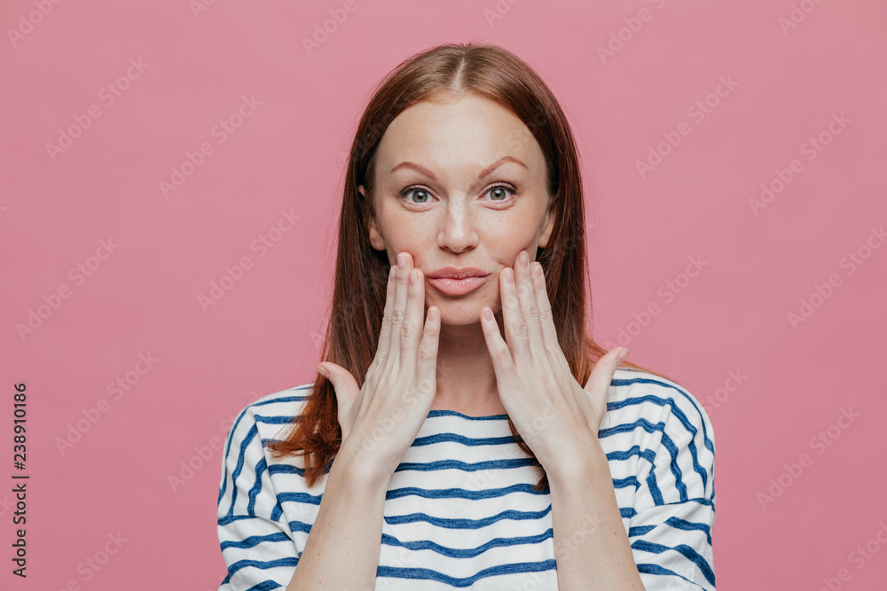 Headshot of freckled pretty woman keeps hands near lips, has minimal make up, healthy skin, brown hair, demonstrates natural beauty, dressed in striped outfit, isolated over pink background. Headshot of freckled pretty woman keeps hands near lips, has minimal make up, healthy skin, brown hair, demonstrates natural beauty, dressed in striped outfit, isolated over pink background.