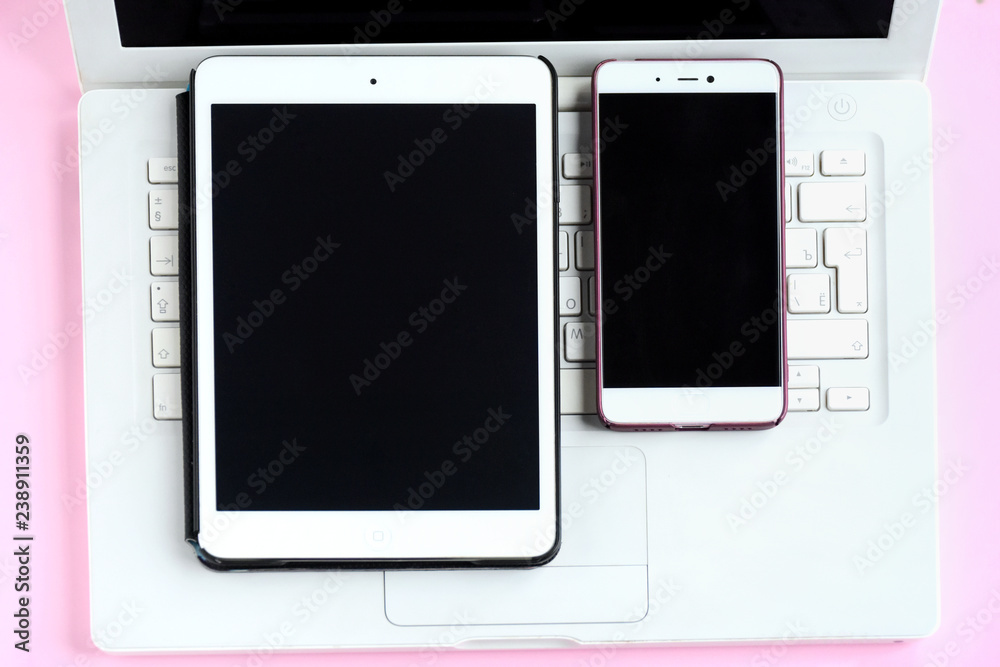 Tablet and mobile phone with black screens on white laptop keyboard. Electronic touch device with selective focus on pink background. Mobility web office with touchscreen Tablet and mobile phone with black screens on white laptop keyboard. Electronic touch device with selective focus on pink background. Mobility web office with touchscreen