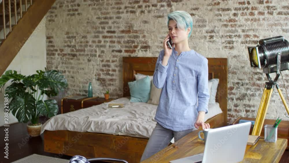 Tilt down shot of young hipster woman with blue hair standing by desk in loft apartment and chatting on phone