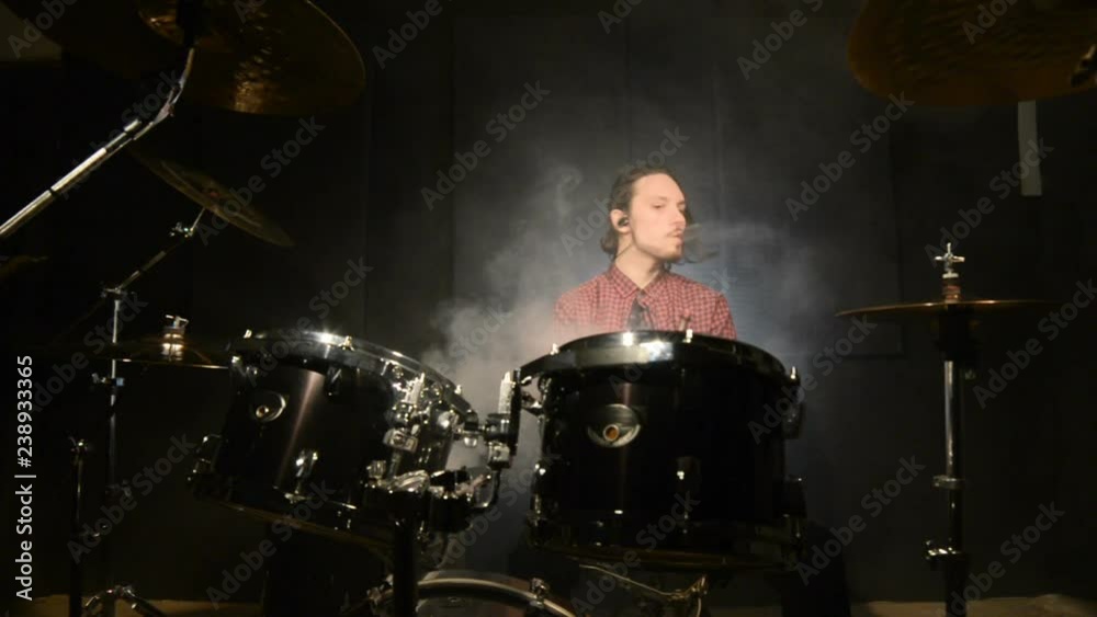 A long-haired drummer sitting behind a drum kit is hovering with a vape. Vape culture in music. Static plan A long-haired drummer sitting behind a drum kit is hovering with a vape. Vape culture in music. Static plan