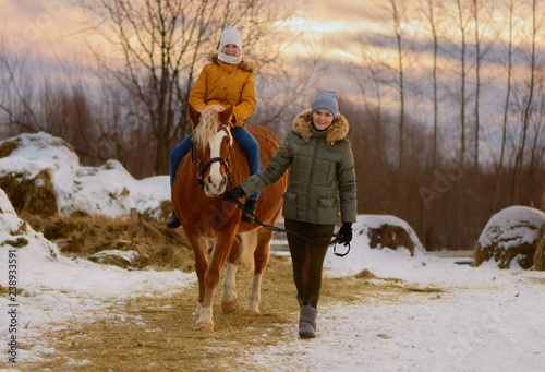 The happy female child is riding a horse. The joyful caucasian woman is holding the reins of her red mare in rural.