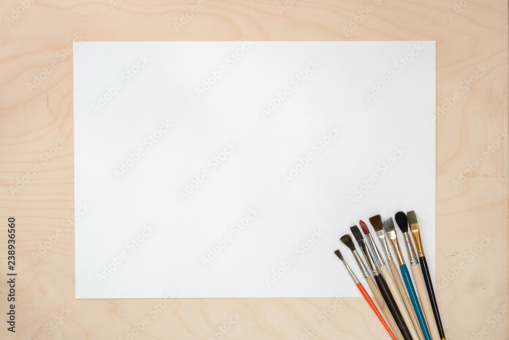 Artist Paint Brushes with Blank White Paper Lying on Wooden Table. Drawing Tools in Art Studio. Top View. Artist Paint Brushes with Blank White Paper Lying on Wooden Table. Drawing Tools in Art Studio. Top View.