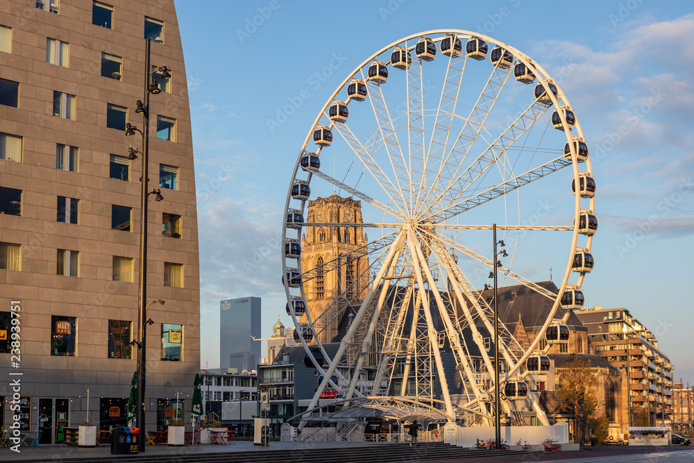 Ferris wheel next to the Markthal in the city centre of Rotterdam, The Netherlands, with behind it a church tower visible through the spokes on a sunny morning with blue sky