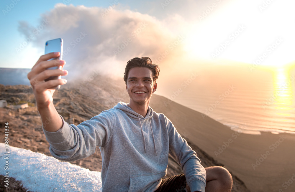 Happy young handsome man with smartphone, making selfie on a mountain hill in sunset light.