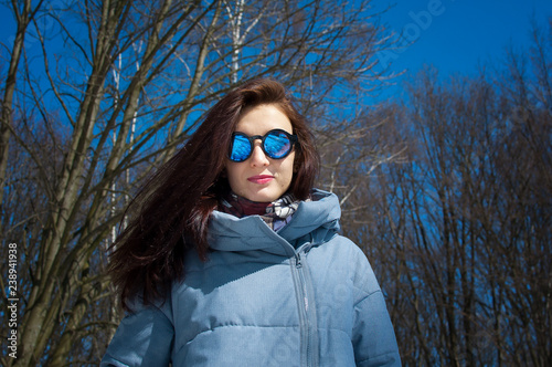 Close portrait of a long-haired brunette in mirrored blue sunglasses and winter clothes against a bright blue sky and bare trees. Winter holidays  waiting for spring
