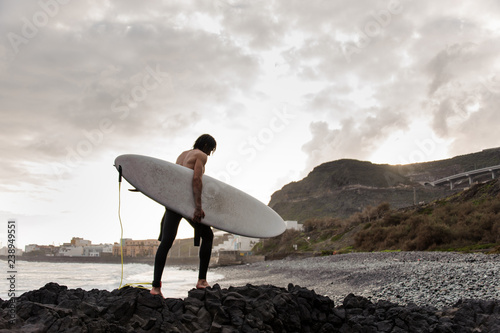 Side view brunette boy walking in the black swimsuit with naked torso with a white surf in his hands on the shore