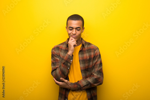Young african american man on vibrant yellow background looking down with the hand on the chin