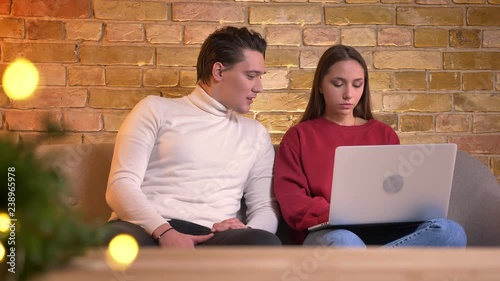 Portrait of cyoung brunette girl working with laptop and laughing with friend sitting next to her on sofa in home atmosphere. photo