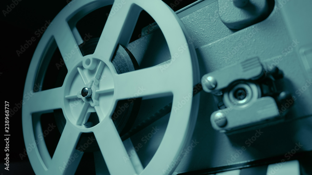 Obraz premium Film projector with dramatic blue lighting and selective focus. Retro film production still life. Concept of film-making. Old 8mm projector is turning off. Close-up of a reel with a film
