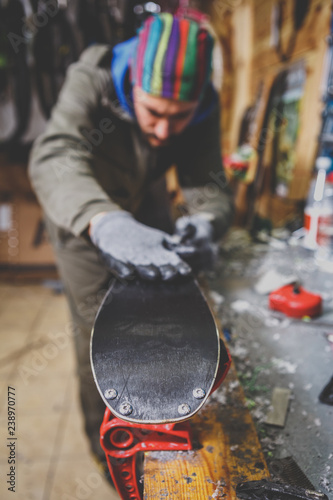 Theme repairs and maintenance of skis. The male worker is repairing work clothes, applying wax on the sliding surface onto skis mounted on ski vise of red color.The concept of service in the workshop