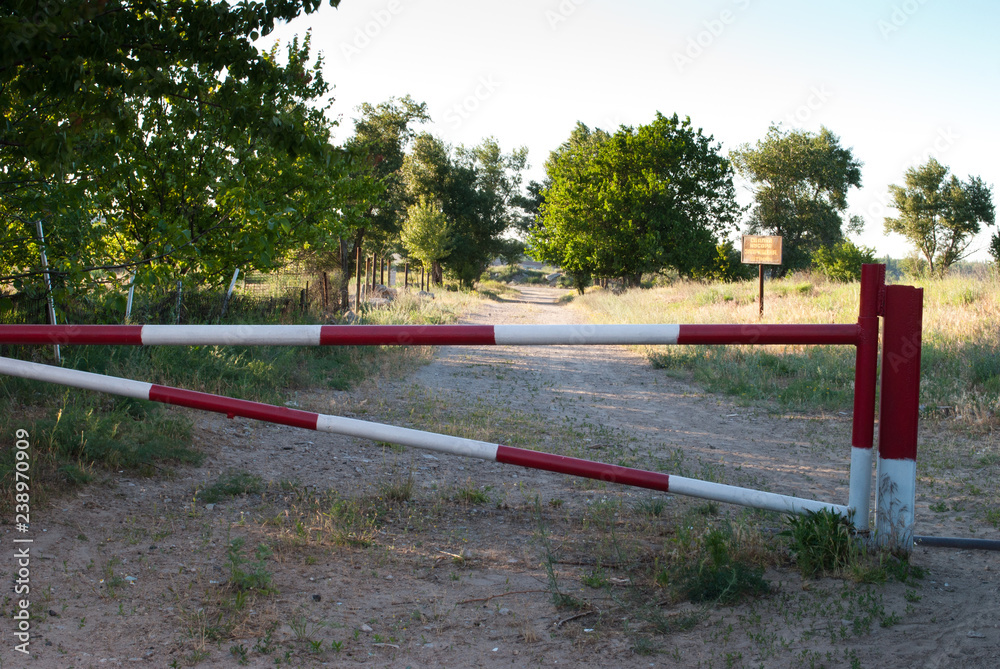 Metal barrier of red and white color, prohibition of travel on the road, against the background of a path of green trees and grass, spring rest way Metal barrier of red and white color, prohibition of travel on the road, against the background of a path of green trees and grass, spring rest way