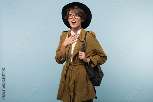 Young surprised woman with dark short hair in trench coat,eyeglasses and hat with backpack amazedly looking aside over blue background isolated photo