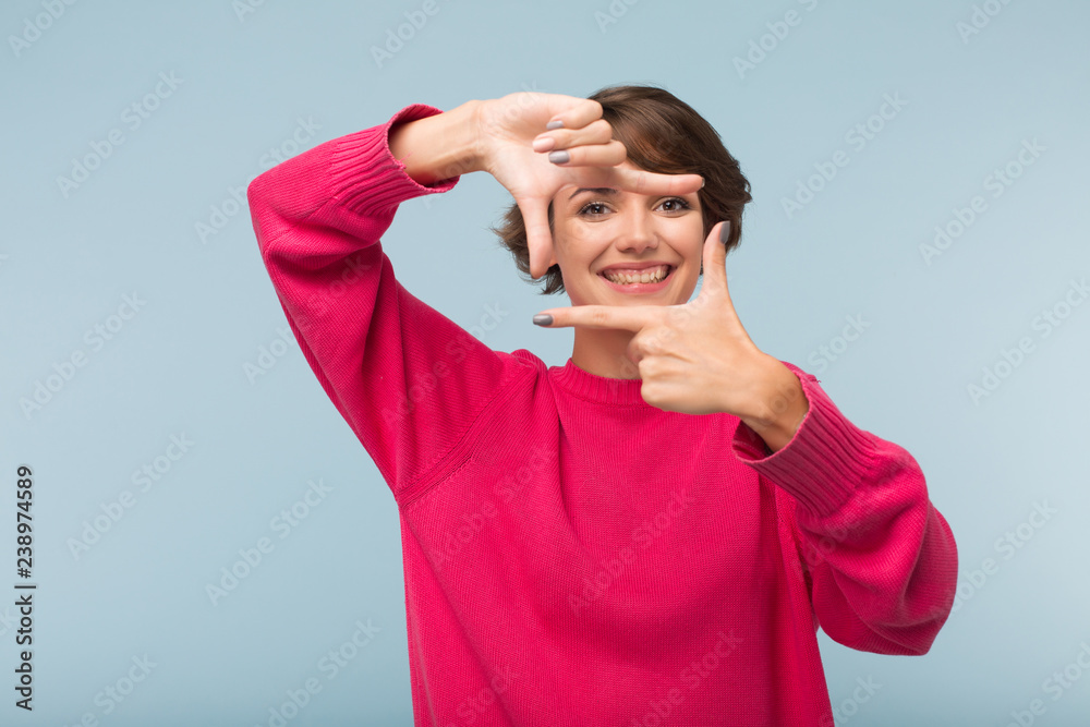 Pretty smiling girl in pink sweater making frame with fingers while happily looking in camera over blue background Pretty smiling girl in pink sweater making frame with fingers while happily looking in camera over blue background
