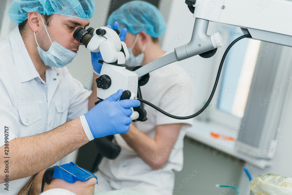 Doctor used microscope. Dentist is treating patient in modern dental office. Operation is carried out using cofferdam. Client is inserted and restored teeth, make denture. Orthodontist and assistant Doctor used microscope. Dentist is treating patient in modern dental office. Operation is carried out using cofferdam. Client is inserted and restored teeth, make denture. Orthodontist and assistant