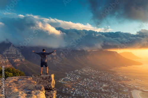 Young man standing on the edge at the top of Lion's head mountain in Cape Town with a beautiful sunset view