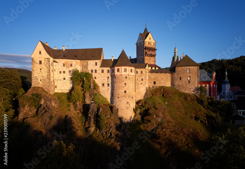 Aerial view on Loket Castle, Burg Elbogen, gothic style castle on a big rock against green spruce forest, massive fortification illuminated by setting sun. Close to Karlovy vary, Czech republic