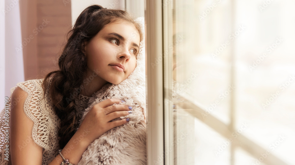 beautiful brunette girl sitting on the windowsill hugging a soft beige pillow and looking out the window beautiful brunette girl sitting on the windowsill hugging a soft beige pillow and looking out the window
