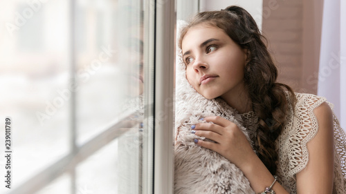 beautiful brunette girl sitting on the windowsill hugging a soft beige pillow and looking out the window