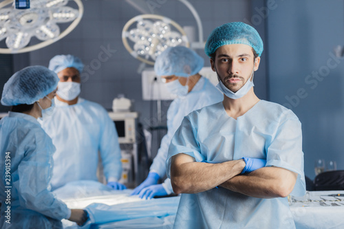 Portrait of handsome caucasian bearded young surgeon looking at camera with colleagues on background while standing in surgery with folded arms.