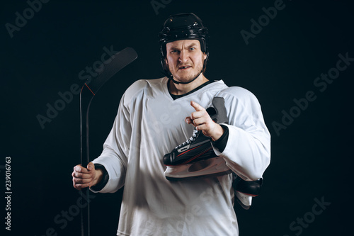 Portrait of a professional hockey player in a protective sportswear and helmet posing with hockey stick and one skate in hands on a black background.