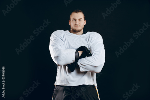 Portrait of ice-hockey player in white headgear and defensive suit looking at camera with folding crossed hands against black background.