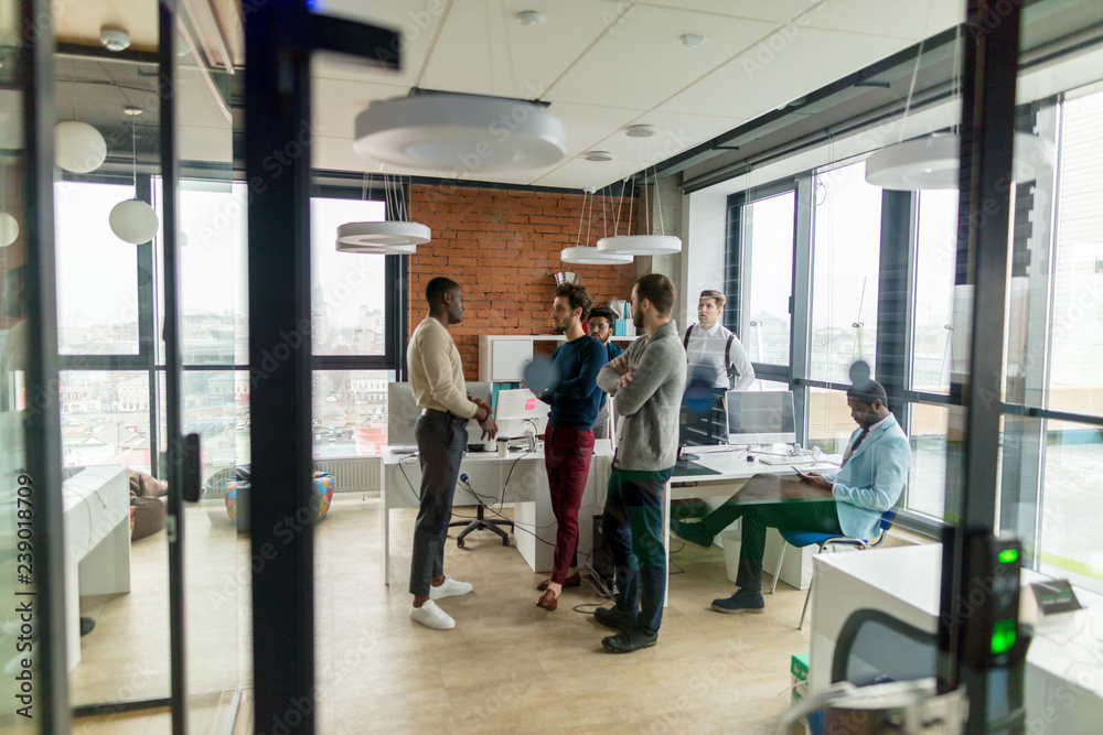 Long distance picture of multiracial male creative team of software developers discussing work agenda in modern city office with panoramic window. Concept of business development.