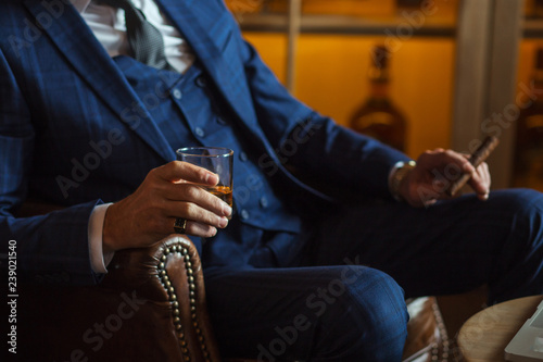 Well-dressed unrecognizable caucasian man in expensive tailored suit relaxing with cigar and alcohol, sitting at his office in leather chair