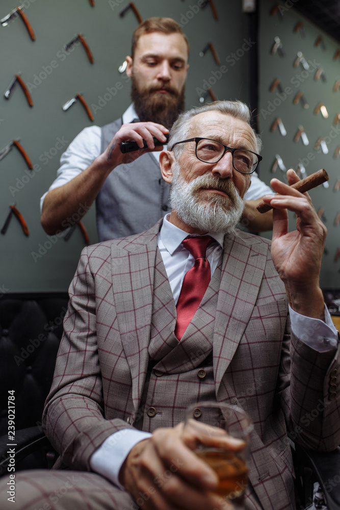 Φωτογραφία Professional young barber giving a haircut with concentration to a rich respectable gentleman, standing and holding razor while relaxed elderly client is smoking cigar and drinking alcohol