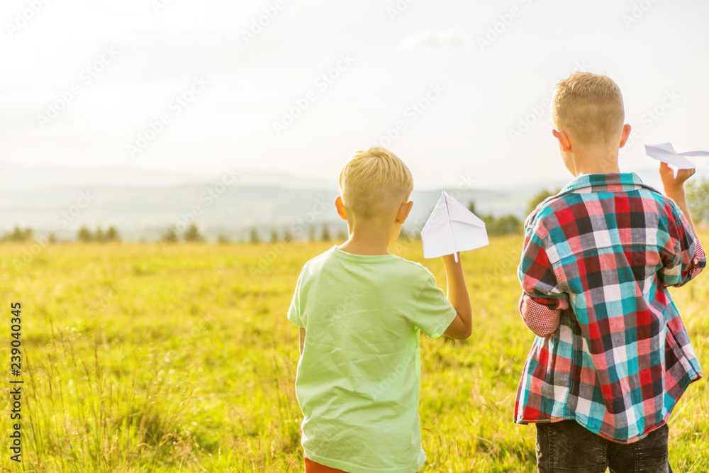 two blonde boys are playing with paper airplanes in a meadow, having fun two blonde boys are playing with paper airplanes in a meadow, having fun