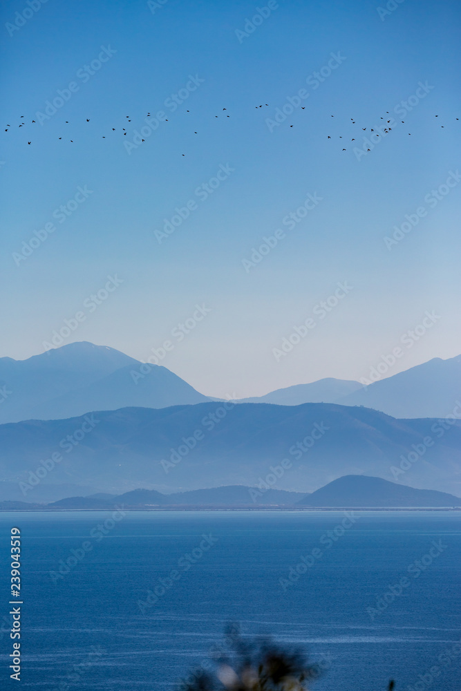Magical vertical view. blue sea water, misty layered mountain range and flying flock of birds in the clear blue sky