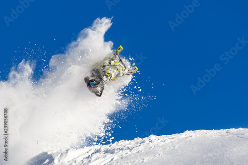 rider on a snowmobile in a jump in the air upside down. from left to right, long trail of big snow storm. winter mountain valley