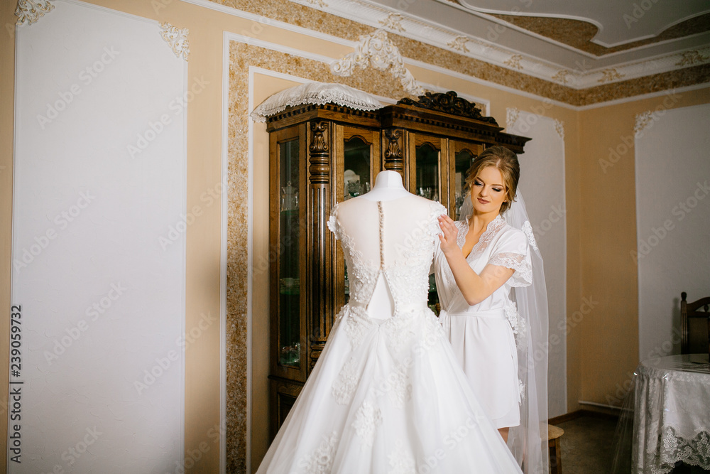 Attractive bride in white robe, long veil standing near the wedding dress on the mannequin on wedding morning. Preparing for the ceremony Attractive bride in white robe, long veil standing near the wedding dress on the mannequin on wedding morning. Preparing for the ceremony