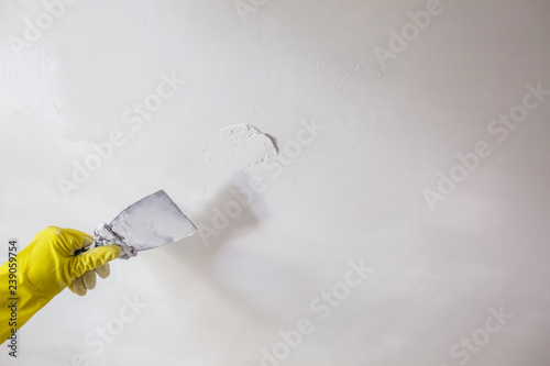 worker's hand in yellow gloves holding putty knife patching a hole with spatula with plaster or putty in white wall. Renovation and repair process, remodeling interior of room at apartment building