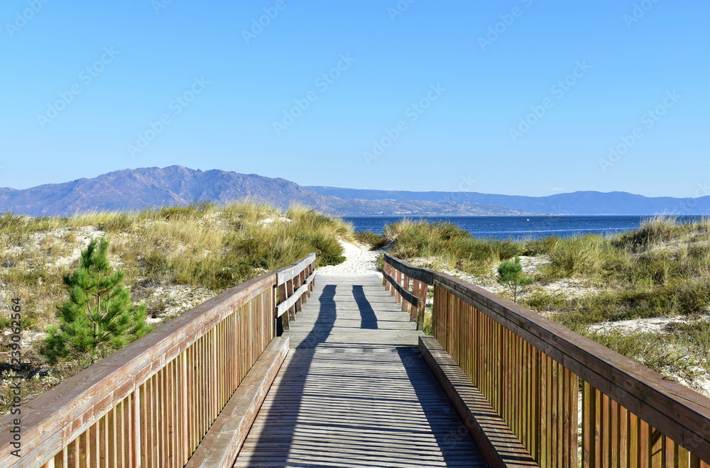 Beach with boardwalk and vegetation in sand dunes. Small mountain and blue sea, clear sky. Sunny day, Galicia, Spain.