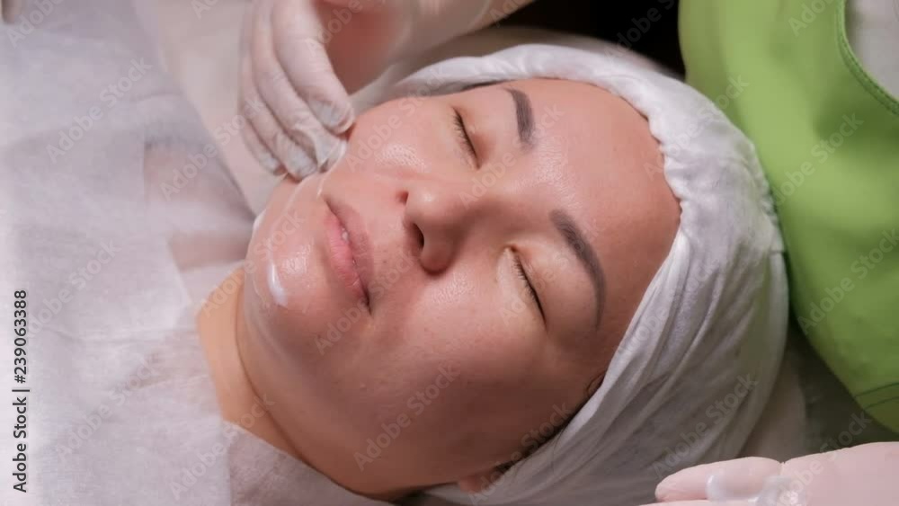 Professional beautician in white gloves puts a nourishing cream on the face of an Asian woman. Cosmetic procedure in the beauty salon. Cleaning and rejuvenation of the skin in the Cosmetology Center. Professional beautician in white gloves puts a nourishing cream on the face of an Asian woman. Cosmetic procedure in the beauty salon. Cleaning and rejuvenation of the skin in the Cosmetology Center.