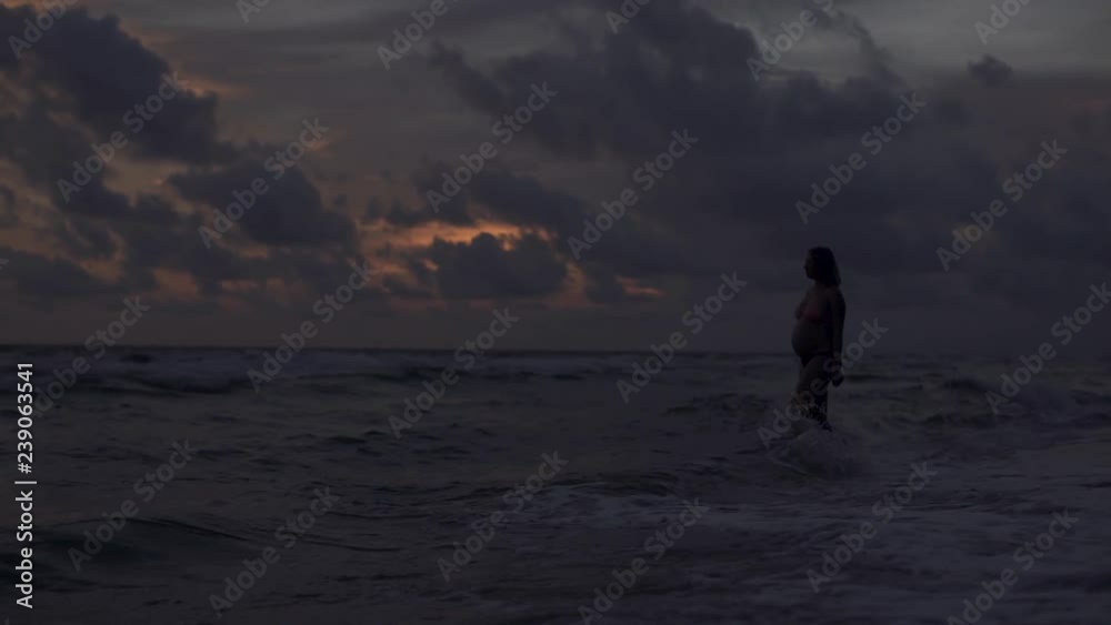 A young pregnant blonde woman walks along the ocean at sunset 4k