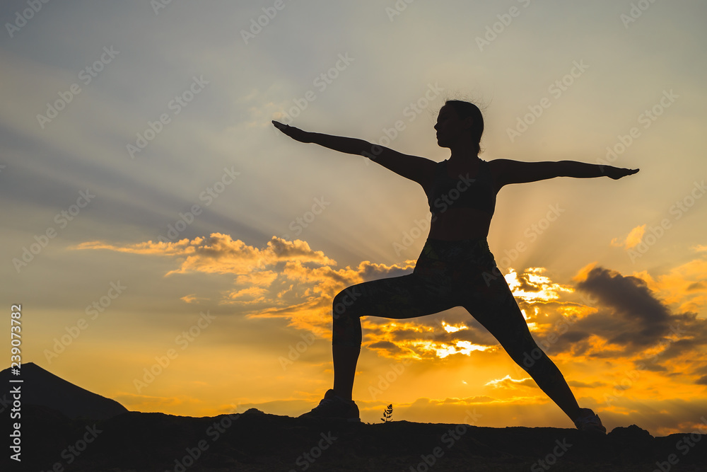 Silhouette of young woman practicing yoga or pilates at sunset or sunrise in beautiful mountain location.