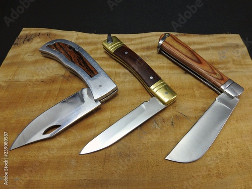 Three folding pocket knives with the half-opened blade, on a wooden surface. Different types of handle materials and colors. Black background.