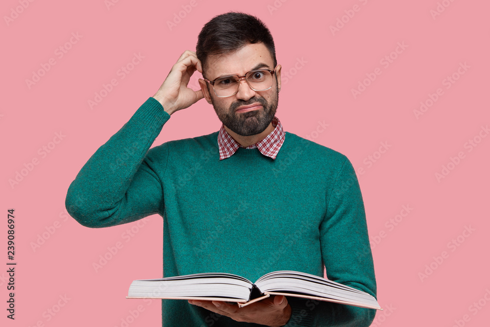 Dissatisfied Caucasian man with thick bristle, scratches head in bewilderment, wears spectacles and green sweater, holds textbook, isolated over pink background. Clever male reader poses indoor Dissatisfied Caucasian man with thick bristle, scratches head in bewilderment, wears spectacles and green sweater, holds textbook, isolated over pink background. Clever male reader poses indoor