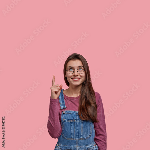 Vertical shot of attractive dark haired smiling woman looks joyfully upwards  points with index finger at ceiling  notices something pleasant  dressed in denim overalls  isolated over pink background