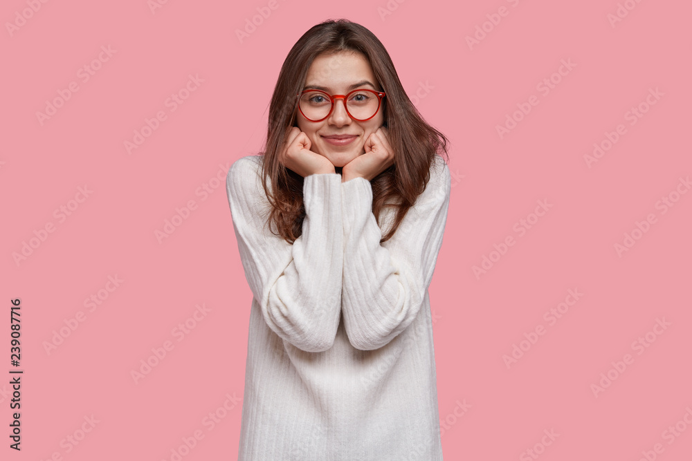 Delighted happy woman holds chin with both hands, wears red rim spectacles and long white sweater, has gentle smile on face, isolated over pink background, being always optimistic and pleased Delighted happy woman holds chin with both hands, wears red rim spectacles and long white sweater, has gentle smile on face, isolated over pink background, being always optimistic and pleased