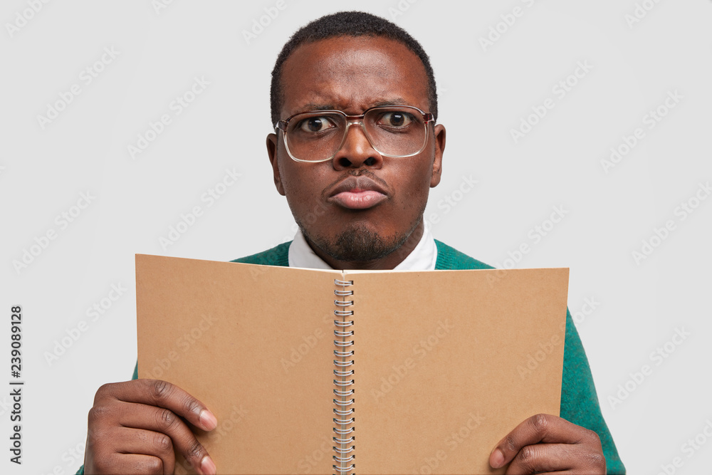 Close up shot of displeased young African American man with gloomy facial expression, purses lips, holds spiral notepad, wears big spectacles, isolated over white wall. Male nerd studies indoor Close up shot of displeased young African American man with gloomy facial expression, purses lips, holds spiral notepad, wears big spectacles, isolated over white wall. Male nerd studies indoor