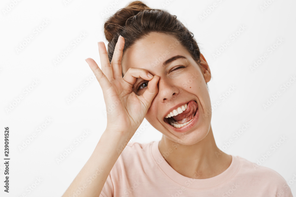 Headshot of entusiastic funny charismatic woman fooling around making funny faces sticking out tongue joyfully as tilting head showing okay gesture over eye, peeking playfully at camera Headshot of entusiastic funny charismatic woman fooling around making funny faces sticking out tongue joyfully as tilting head showing okay gesture over eye, peeking playfully at camera