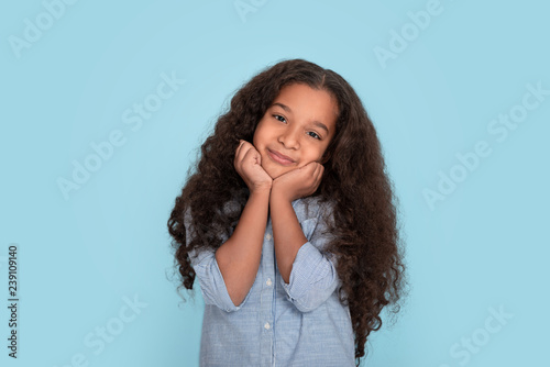 Close up emotional portrait of young frizzy smiling girl wearing blue shirt on blue background in studio. She holds her cheeks and smiles photo