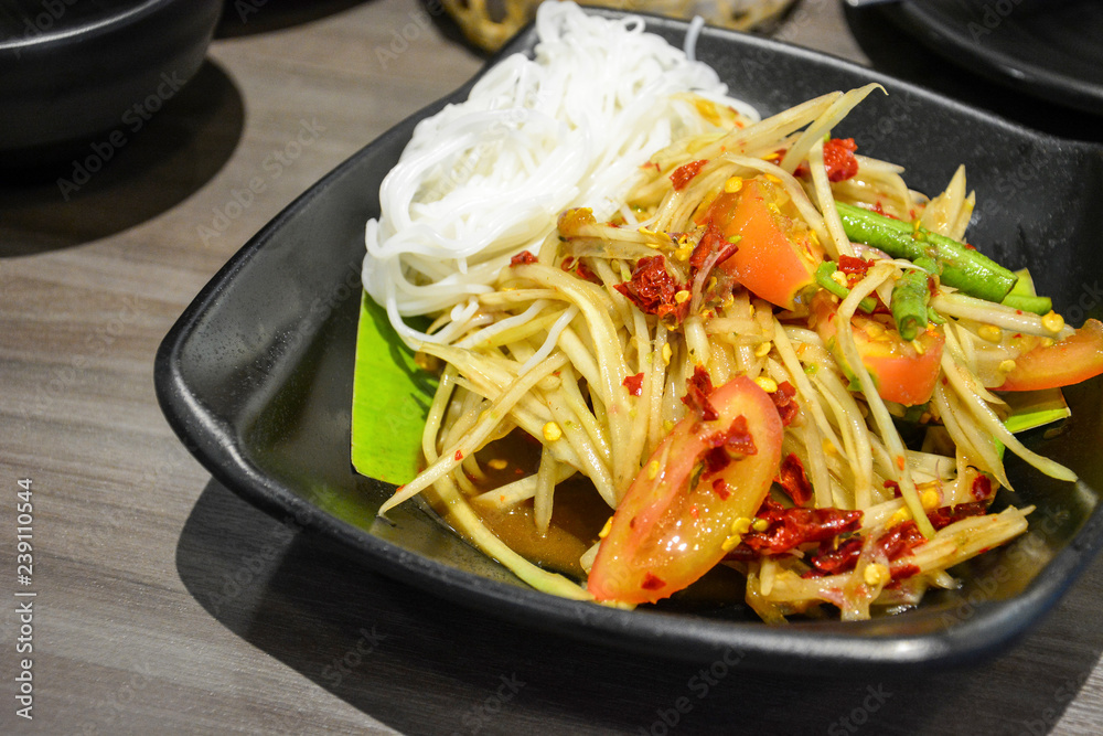 famous and popular Thai street food, green papaya spicy salad with pickled fish or Som Tum served with Thai rice noodles in black plate on wooden table background famous and popular Thai street food, green papaya spicy salad with pickled fish or Som Tum served with Thai rice noodles in black plate on wooden table background
