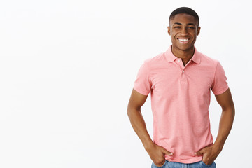 Waist-up shot of attractive positive and happy young african american male entrepreneur in pink polo shirt holding hands in pockets and smiling with self-assured delighted grin over white background