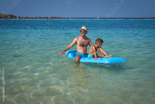 Attractive European man with his son is sitting on a blue air mattress on the beach. They are relaxing and enjoying their holidays.