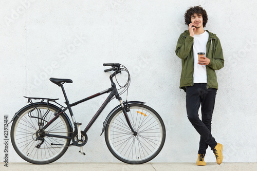 Fototapeta Naklejka Na Ścianę i Meble -  Handsome man with curly hair, dressed in green anorak, black trousers, has curly hair, speaks with girlfriend via cell phone, carries takeaway coffee, poses against white background near his bicycle