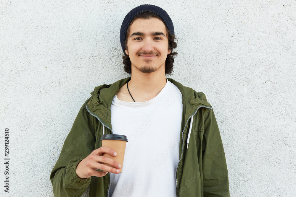 Headshot of pleased hipster guy holds takeaway coffee, dressed in white t shirt and green jacket, enjoys spare time, wears black hat, models over white background, has curly hair. People and drinking Headshot of pleased hipster guy holds takeaway coffee, dressed in white t shirt and green jacket, enjoys spare time, wears black hat, models over white background, has curly hair. People and drinking