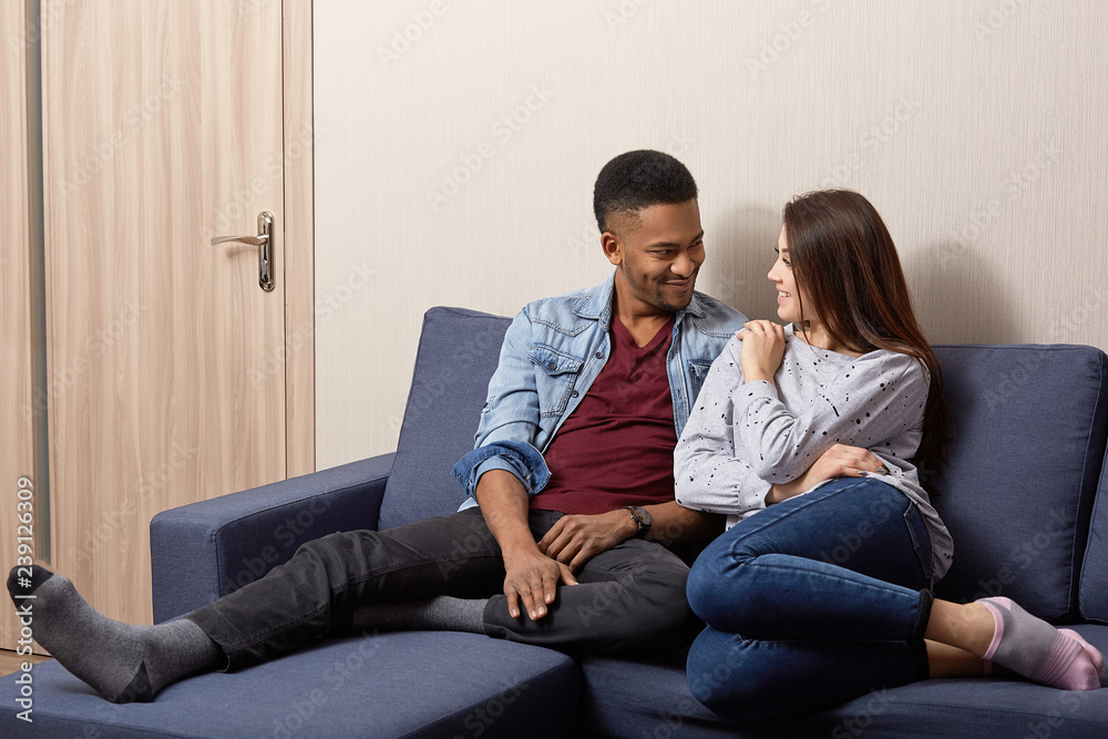 Pleased dark skinned man and his Caucasian girlfriend look at each other with love, pose on comfortable couch, enjoy pleasant talk, coziness, spend weekend at home. Interracial family couple. Pleased dark skinned man and his Caucasian girlfriend look at each other with love, pose on comfortable couch, enjoy pleasant talk, coziness, spend weekend at home. Interracial family couple.